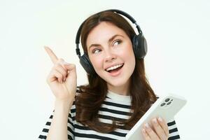 Happy beautiful woman, listening music in wireless headphones, holding mobile phone, pointing left at copy space, showing advertisement, white background photo