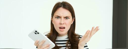 Portrait of angry, confused young woman shrugging shoulders while using mobile phone, holding smartphone with annoyed face expression, frowning, standing over white background photo