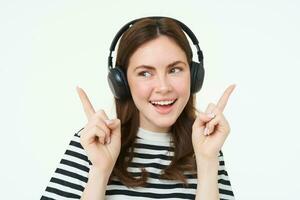 Dancing girl in wireless headphones pointing at empty space for music store promo text, showing advertisement, isolated over white background photo