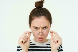 Portrait of shocked, angry woman frowning, looks confused at camera, takes off glasses with frustrated face expression, stands over white background photo