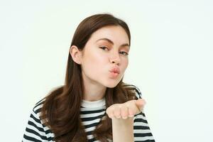 Portrait of cute girl sending you, blowing air kiss, holding palm near puckered lips, smiling, standing over white background photo