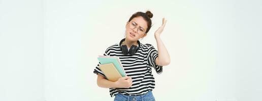 Image of upset young woman, student in glasses complains at difficult task at university, holding notebooks, forgot to do something, white background photo