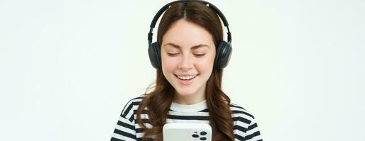 Image of young woman in headphones, using smartphone and laughing, watching video on mobile phone, listens to music on streaming service app, white background photo
