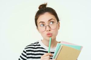 cerca arriba retrato de niña en anteojos, pensamiento, participación cuaderno y bolígrafo, mirando arriba y reflexionando, haciendo decisión, en pie terminado blanco antecedentes foto