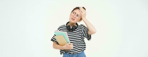 Portrait of compllicated, sad young woman in glasses, carries homework notebooks, facepalms, looks disappointed, stands over white background photo