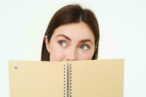 Young woman holds notebook daily planner next to her face, writing down homework, making notes, looking thoughtful, standing over white background photo