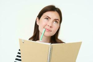 Portrait of student doing her homework, holding notebook and pen, thinking while making notes in her notebook, standing over white background photo