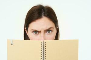 Portrait of woman with frowning face, holding notebook, organizer for notes and memos, looking suspicious and doubtful at camera, standing over white background photo