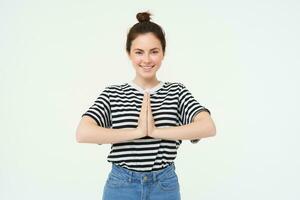 Portrait of beautiful young woman, holds hands together, namaste gesture, say thank you, expresses gratitude, appreciation, stands over white background photo