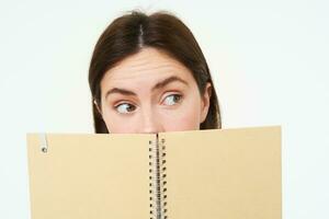 Close up portrait of woman with thoughtful expression, looking aside, hiding half of face behind notebook, standing with memo planner over white background photo