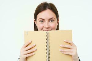 Image of young woman with notebook, holding her personal diary, work planner, smiling, reading something, standing over white background photo