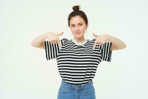 Portrait of confident, smiling brunette woman, pointing fingers down, showing banner on bottom, follow link below gesture, standing over white background photo