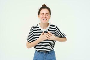 Portrait of happy, smiling young woman stands with eyes closed, holds hands on heart, laughs, remember something funny, isolated over white background photo