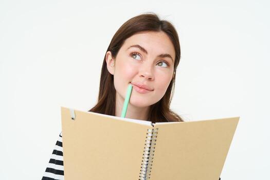 retrato de creativo joven mujer, mirando considerado, participación bolígrafo y computadora portátil, escritura en su planificador, pensando mientras haciendo notas, en pie terminado blanco antecedentes foto