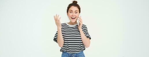 Portrait of excited woman answers phone call, reacts amazed to wonderful news received over the telephone, standing over white background photo