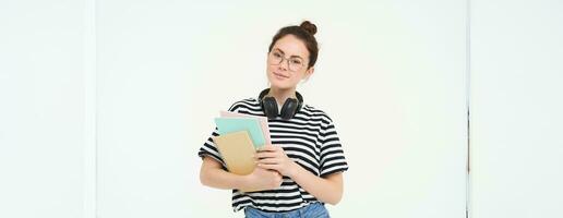 Student and education concept. Young woman with books, notes and pen standing over white background, college girl with headphones over neck posing in studio photo