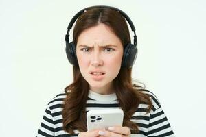 Image of puzzled young woman in headphones, holds smartphone, frowning and looking confused at camera, standing over white background photo