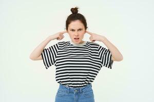 Image of annoyed, bothered woman plugs, shuts her ears with fingers, disturbed by loud noise, stands over white background photo