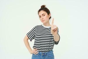 Image of confident woman, shows one finger, taboo, stop gesture, disapprove smth, gives warning, stands over white background photo