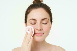 Image of young woman taking off her makeup with cotton pads, using facial cleanser, cleaning her face with skincare treatment, standing over white background photo