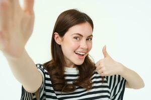 Portrait of smiling woman recommending product, shows thumbs up, takes selfie with something good, copy space, standing isolated on white background photo