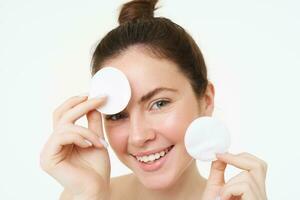 Beauty and cosmetics. Young smiling woman washing her face with cotton pads, using facial cleanser, looking happy at camera photo