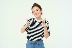 Portrait of cute girl, student shows thumbs up, recommends product, approves choice, likes item, stands over white background photo