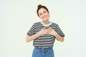 Image of beautiful woman in casual clothes, holds hands on heart and smiles, express gratitude, being thankful, stands over white background photo