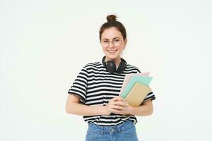 Image of young woman, tutor with books and notebooks, wearing headphones over her neck, isolated on white background. Student lifestyle concept photo