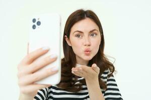 Portrait of young european woman taking selfie on smartphone, holding white mobile phone and posing for photos, isolated against white background photo