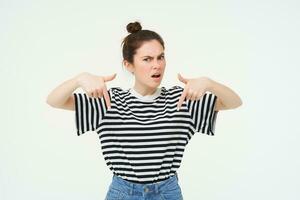 Frustrated girl points down, frowns and looks angry, asks questions with upset face expression, stands over white background photo