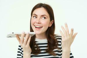 Image of cute brunette woman talking into speakerphone, holding mobile phone near mouth, records her voice, sends a voicemessage, using online translator app, white background photo