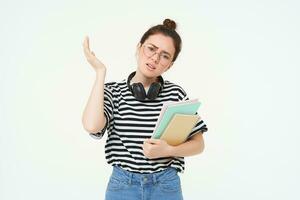 Portrait of compllicated, sad young woman in glasses, carries homework notebooks, facepalms, looks disappointed, stands over white background photo