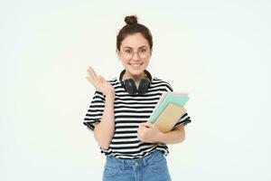 Portrait of girl in glasses, with headphones over her neck, holds notebooks and study material for university or college, looks happy and surprised, white background photo