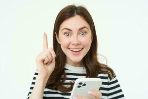 Got an idea. Smiling woman, raises her finger and holds smartphone, suggests something, makes eureka gesture, has a solution, white background photo