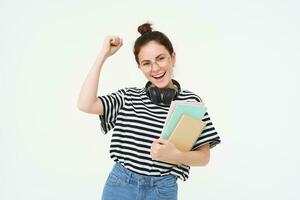 Portrait of young woman in glasses, wearing headphones over neck, holding planner, notebooks and study material, cheering, rooting for you, celebrating, standing over white background photo