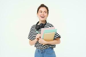 Student and education concept. Young woman with books, notes and pen standing over white background, college girl with headphones over neck posing in studio photo