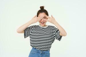 Happy young woman, bday girl peeking through fingers, holds hands on eyes, waiting for surprise, stands over white background photo