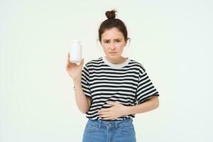 Woman takes painkillers, pills from stomach ache, menstrual pain, holds hand on belly, shows bottle with tablets, stands over white background photo