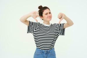 Young woman gives negative feedback, shows thumbs down, disapproves product, dislikes smth bad, stands over white background photo