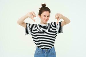 retrato de decepcionado, joven moderno mujer, estudiante muestra pulgares abajo, no le gusta algo, desaprueba, soportes terminado blanco antecedentes foto