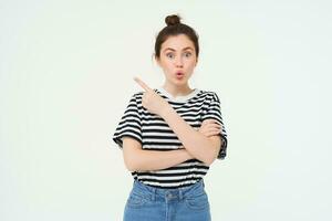 Image of girl with surprised face, looks amazed, points finger left, shows advertisement, white background photo