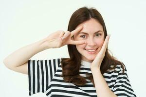 Portrait of beautiful, smiling female model, touches her clear glowing skin, shows peace sign and looks happy at camera, isolate on white background photo