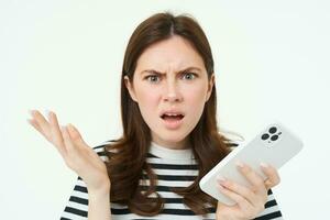 Portrait of angry, confused young woman shrugging shoulders while using mobile phone, holding smartphone with annoyed face expression, frowning, standing over white background photo