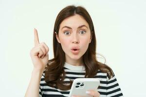 Portrait of excited woman raising finger, eureka gesture, has an idea or solution, holds mobile phone, white background photo
