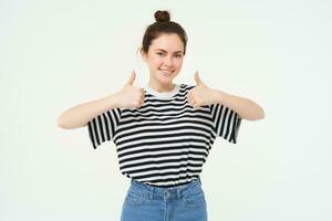 Portrait of cute girl, student shows thumbs up, recommends product, approves choice, likes item, stands over white background photo