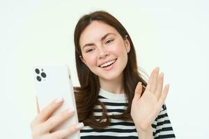 Happy young woman waves her hand at smartphone camera, saying hello to viewers on social media app, video chats on mobile phone app, white background photo