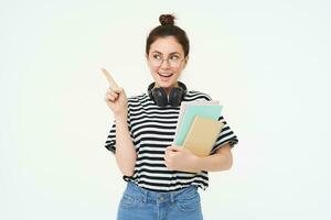 Image of young modern girl, student or teacher in glasses, holding documents and notebooks, pointing at upper left corner with pleased smile, showing advertisement, white background photo