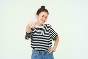 Image of confident, happy young woman in casual clothes, pointing finger at camera, laughing and smiling, standing over white background photo