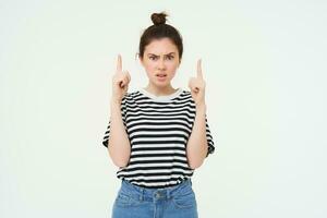 Portrait of shocked woman, raise eyebrows and points fingers up, looks complicated, puzzled by something, standing over white background photo